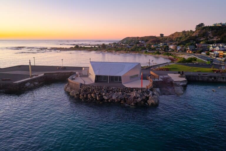 Modern coastal building with curved metal roof on rocky outcrop at sunrise, Kaikōura residential development by LMC Building.